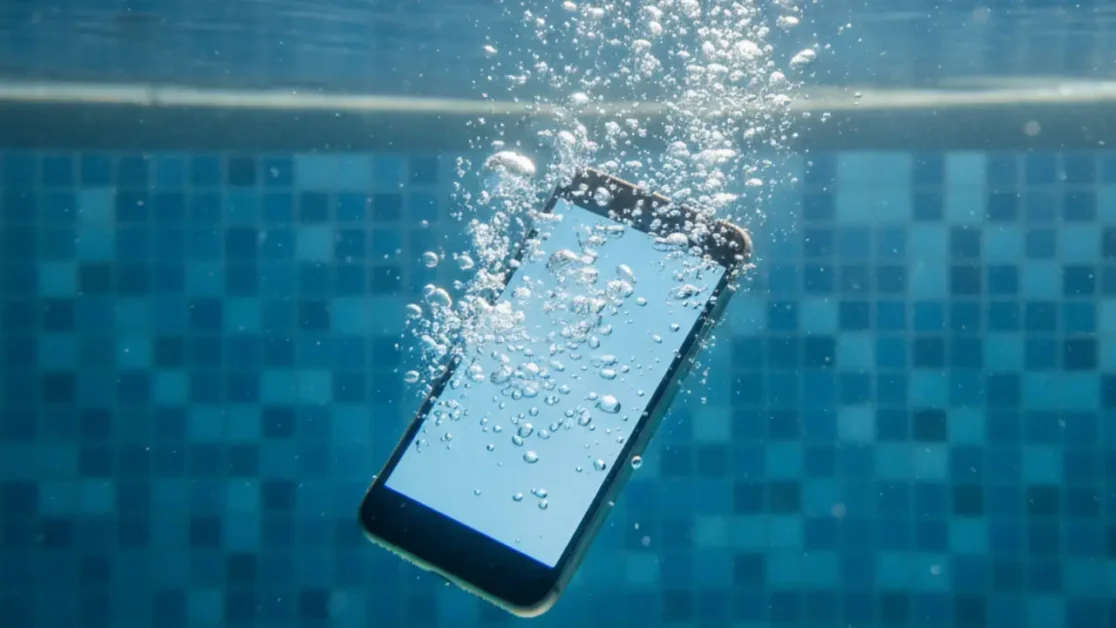 Imagem subaquática de um smartphone afundando lentamente em uma piscina de água cristalina. A tela do aparelho está acesa, emitindo um brilho azulado, enquanto um rastro de bolhas sobe em direção à superfície.